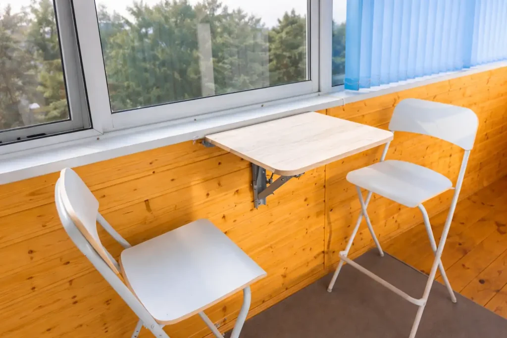 Space-saving enclosed balcony with wall-mounted foldable table and white chairs for compact apartment living