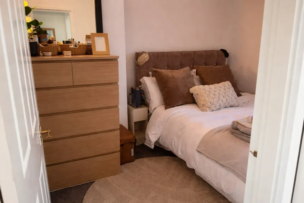 Small cozy bedroom viewed from doorway with wooden dresser, soft bedding, and neutral decor