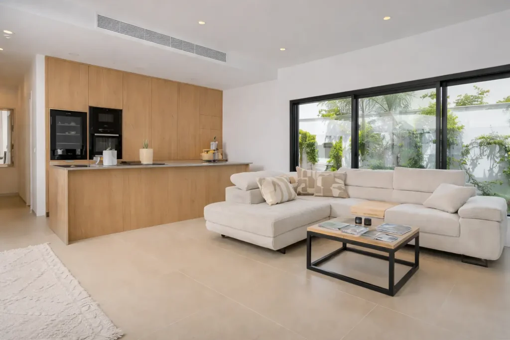 Modern open-plan living room with beige sectional sofa, wooden kitchen cabinetry, black coffee table, and large sliding glass doors with garden view